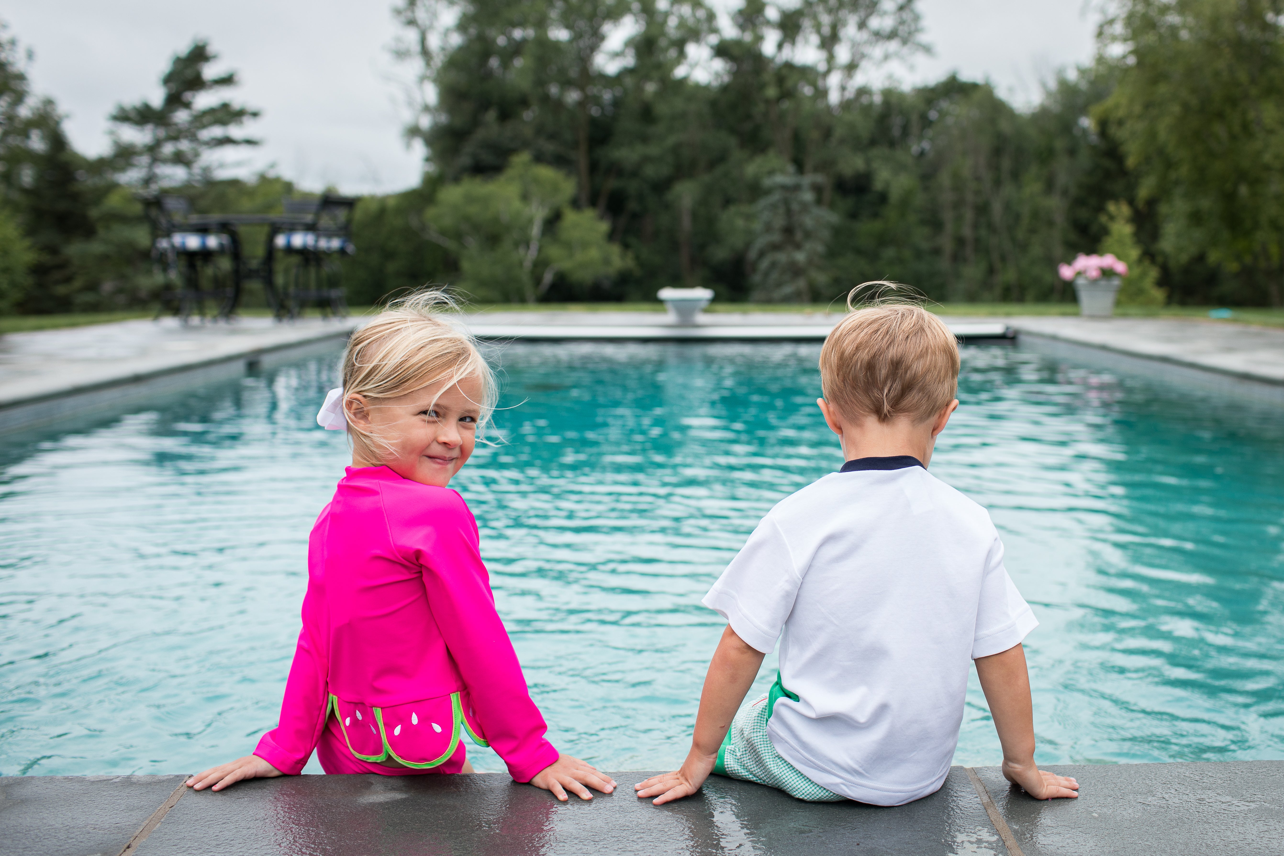 a young boy and girl by the pool, the girl, in a pink rashguard, is looking back and smiling